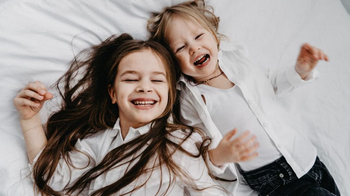 Two happy sisters with long hair laughing together while lying on a bed.