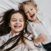 Two happy sisters with long hair laughing together while lying on a bed.